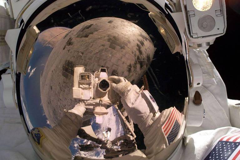 Reflexo de astronauta segurando câmera e trabalhando no espaço é visível na viseira do capacete
