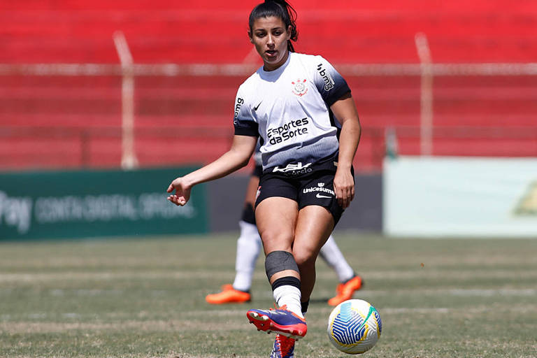 Uma jogadora de futebol está em campo, prestes a chutar a bola. Ela veste um uniforme predominantemente branco com detalhes em preto e está usando meias e caneleiras. O fundo mostra arquibancadas vermelhas e uma grama bem cuidada. A jogadora parece concentrada e em movimento.
