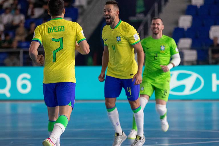 Três jogadores de futsal da seleção brasileira estão em um momento de celebração em quadra. O jogador à esquerda, com a camisa amarela e o número 7, é Dyego. O jogador central, também com a camisa amarela e o número 4, está sorrindo e levantando a mão. O terceiro jogador, à direita, usa uma camisa verde e parece estar se juntando à celebração. O fundo mostra uma quadra de futsal com uma iluminação suave e uma tela publicitária.