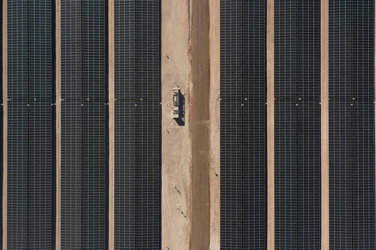 A imagem mostra uma série de painéis solares dispostos verticalmente em uma estrutura de madeira. Os painéis são de cor escura e estão alinhados em fileiras, com uma separação visível entre eles. No centro, há uma peça de metal que parece ser um suporte ou conector.