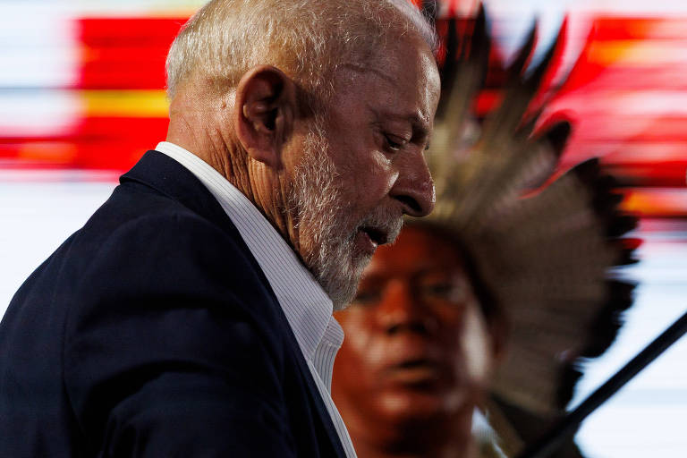 A imagem mostra um homem de cabelo grisalho e barba, vestindo um terno escuro, com uma expressão séria. Ao fundo, há uma pessoa com um cocar indígena, que observa atentamente. O fundo é colorido, com tons de vermelho e amarelo, sugerindo um ambiente de evento.