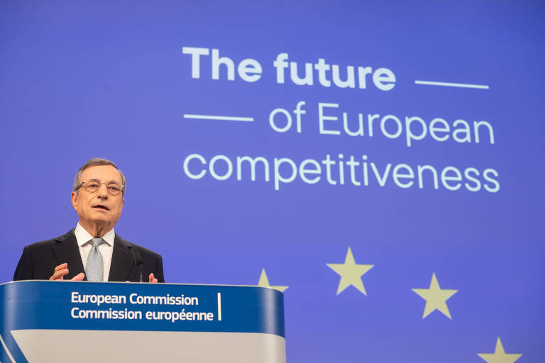 Homem usando terno está em pé atrás de púlpito em discurso em evento. Há um painel azul com a bandeira da União Europeia e a frase "The future of European competitiveness"