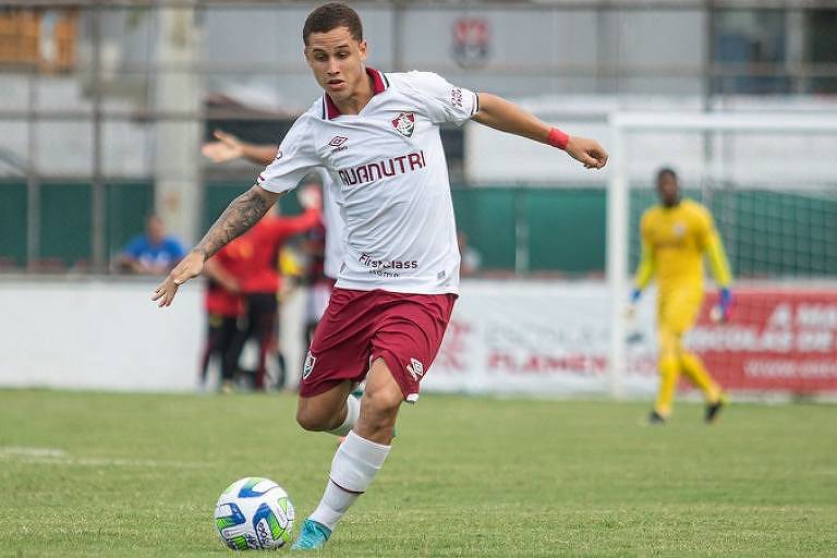 Um jogador de futebol está em movimento, chutando uma bola em um campo. Ele veste um uniforme branco com detalhes em vermelho e o logo de um clube. Ao fundo, há outros jogadores e um goleiro vestido de amarelo. O campo é gramado e há uma rede de gol visível.