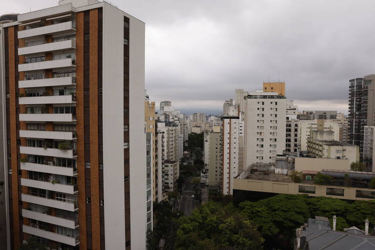 imagem mostra vista de cidade com vários prédios à esquerda e à direita. ao centro, na parte inferior, árvores. O céu está nublado