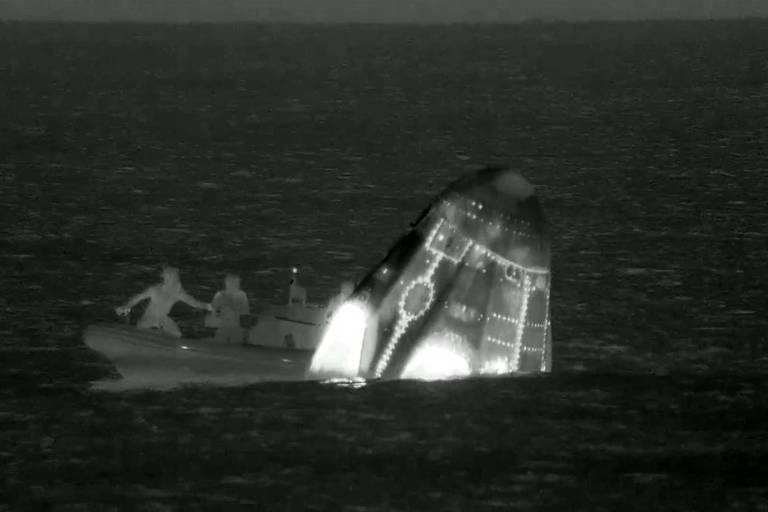 A imagem mostra uma nave espacial parcialmente submersa no oceano, com a parte superior visível e iluminada. Ao lado, há um pequeno barco com pessoas, que parecem estar se aproximando da nave. A cena é capturada em um tom escuro, sugerindo que foi tirada à noite.