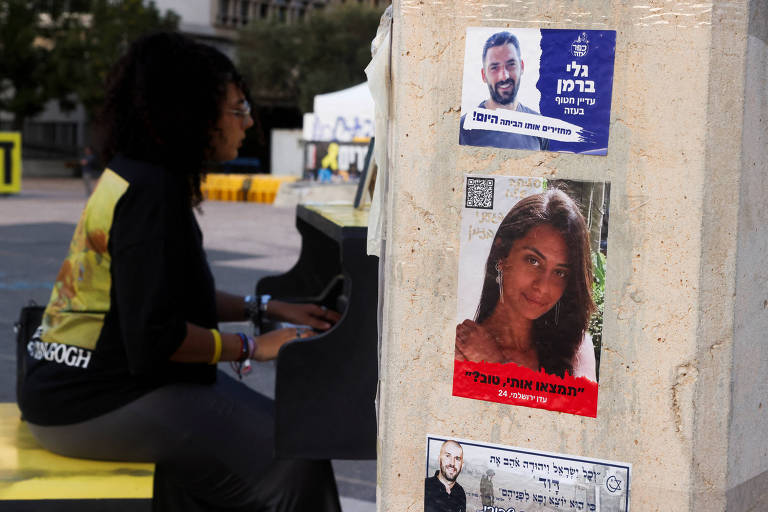 Uma mulher com roupa preta e detalhe em amarelo nas costas é vista de perfil tocando piano ao lado de imagens de pessoas que foram feitas reféns pelo Hamas
