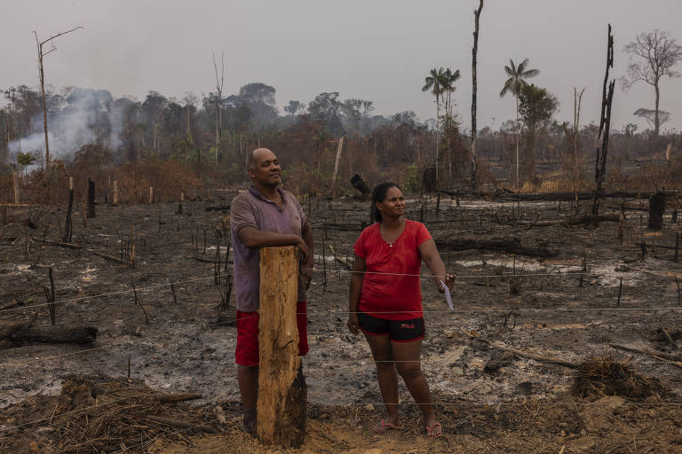 Casal de pessoas negras vestidas de forma simples está encostado a uma cerca, dentro de uma propriedade que parece ter sido devastada por um incêndio recentemente