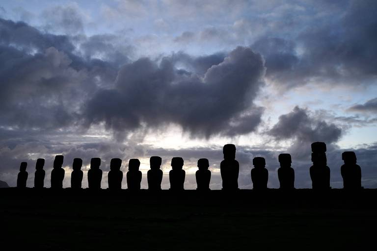 A imagem mostra uma fileira de estátuas Moai em silhouette contra um céu nublado ao entardecer. As estátuas estão alinhadas em uma colina, com nuvens escuras e uma luz suave ao fundo, sugerindo o final do dia.