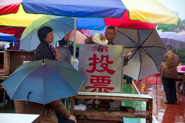 A imagem mostra uma cena de uma feira ao ar livre durante a chuva. Várias pessoas estão abrigadas sob guarda-chuvas coloridos. Um homem idoso está em pé atrás de uma estrutura metálica com um cartaz em vermelho que diz '投票' (votação). Uma mulher com um guarda-chuva azul claro observa ao lado. O chão está molhado devido à chuva.