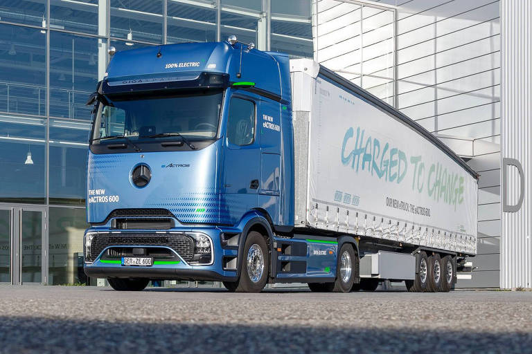 A imagem mostra um caminhão de transporte de cor azul, estacionado em frente a um edifício moderno com grandes janelas de vidro. O caminhão possui uma carroceria branca com detalhes em azul e verde, e está posicionado em uma área pavimentada. O céu está claro, com algumas nuvens visíveis.