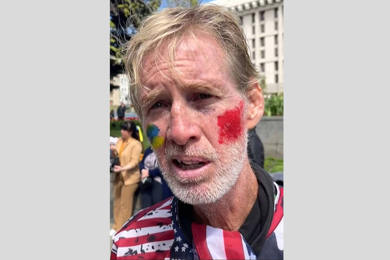 Um homem com cabelo loiro e barba grisalha está em primeiro plano, com pintura facial nas cores azul e amarelo de um lado e vermelho do outro. Ele usa uma camiseta com listras vermelhas e brancas e um padrão de estrelas. Ao fundo, há pessoas e um edifício visível.