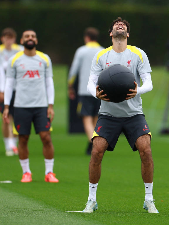 Mohamed Salah (atrás) e Dominik Szoboszlai em treino do Liverpool; os dois sorriem e o segundo olha para cima e segura uma bola da cor preta com as duas mãos