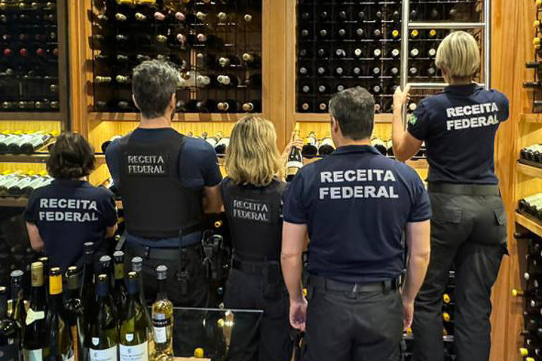 A imagem mostra um ambiente de armazenamento de vinhos, com prateleiras de madeira repletas de garrafas de vinho. Cinco pessoas estão de costas, organizando as garrafas nas prateleiras. O espaço é bem iluminado e possui um piso de madeira.