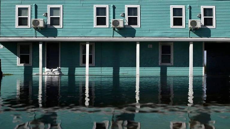 A imagem mostra um edifício de cor azul claro parcialmente submerso em água devido a uma inundação. O nível da água chega até as colunas do prédio, refletindo a estrutura. Há janelas com ar-condicionado visíveis na fachada, e sacos de areia estão empilhados na parte inferior, possivelmente para contenção de água.
