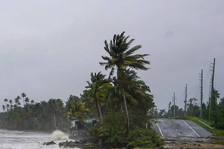 A imagem mostra uma estrada costeira em um dia nublado, com ondas do mar batendo na margem. À esquerda, há palmeiras inclinadas devido ao vento forte. A estrada à direita parece danificada, com algumas partes alagadas. No fundo, há postes de eletricidade alinhados ao longo da estrada.
