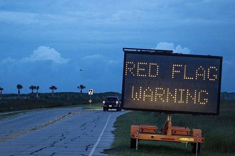 Uma placa eletrônica de aviso está posicionada ao lado de uma estrada, exibindo em inglês a mensagem 'aviso de bandeira vermelha'. O céu está nublado e há algumas palmeiras visíveis ao fundo. Um veículo está se aproximando da placa.
