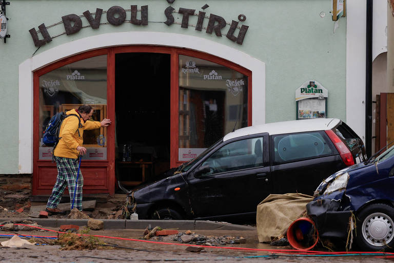 A imagem mostra uma cena de destruição em frente a um restaurante chamado 'U Dvou Štírů'. Um homem com uma jaqueta amarela e calças xadrez caminha em direção ao restaurante, enquanto vários carros danificados estão estacionados na calçada. Um dos carros está parcialmente submerso em lama e detritos. O ambiente parece ter sido afetado por uma enchente, com sujeira e objetos espalhados pelo chão.