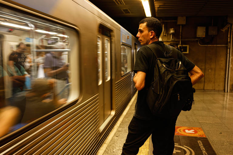 Um homem está de pé na plataforma de uma estação de metrô, olhando para um trem que está se aproximando. Ele usa uma camiseta preta e carrega uma mochila nas costas. Ao fundo, outras pessoas estão visíveis dentro do trem, enquanto a luz da estação ilumina o ambiente.