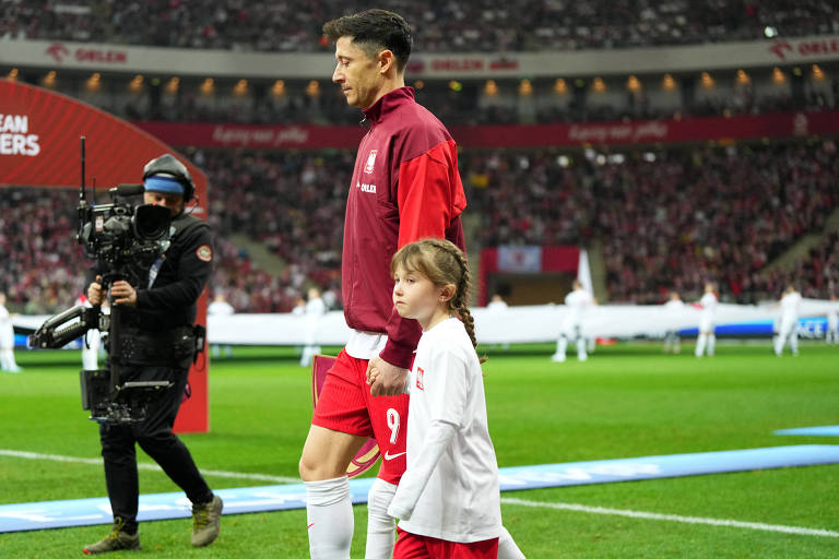 Um jogador de futebol, vestido com um agasalho vermelho e branco, caminha em direção ao campo segurando a mão de uma criança. A criança usa uma camiseta branca com um pequeno emblema. Ao fundo, há uma multidão em um estádio, com bandeiras e uma atmosfera de evento esportivo.
