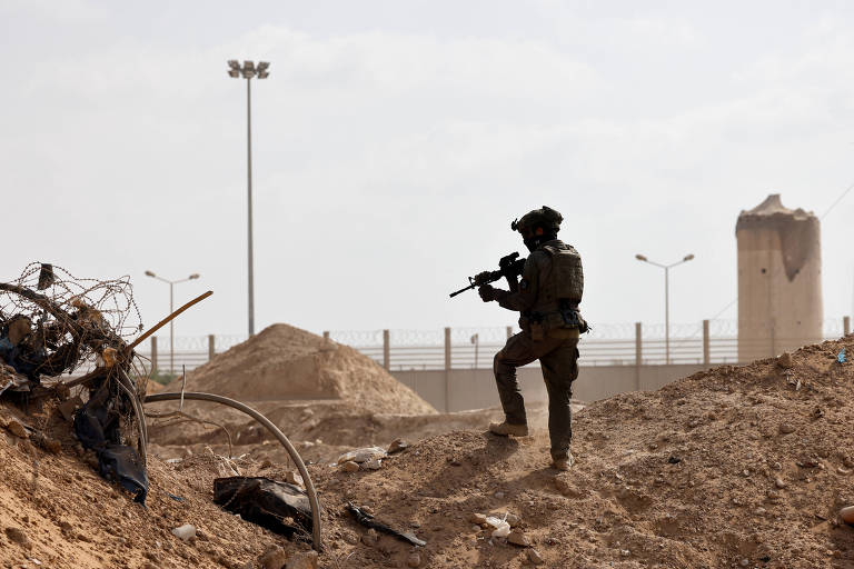 Um soldado armado está posicionado em uma área de conflito, com uma paisagem árida ao fundo. Há montes de terra e destroços ao redor, e estruturas de concreto visíveis ao longe. O céu está nublado, criando uma atmosfera tensa