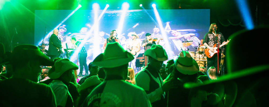 A imagem mostra um show ao vivo com uma banda se apresentando em um palco iluminado por luzes verdes. O fundo exibe uma tela grande com imagens da banda. O público, que usa chapéus, está em primeiro plano, dançando e se divertindo. A atmosfera é animada e festiva.