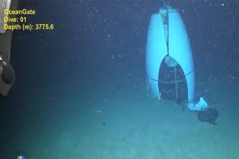 A imagem mostra uma estrutura subaquática, possivelmente uma embarcação ou um objeto semelhante, posicionada no fundo do mar. A estrutura apresenta uma abertura na parte frontal. O ambiente é escuro, com algumas partículas visíveis na água, sugerindo uma profundidade considerável