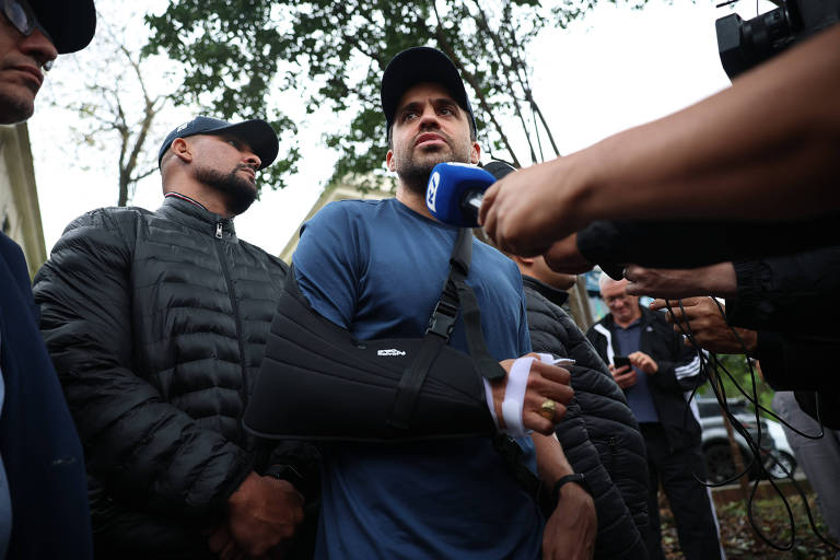 A imagem mostra um homem visto de baixo para cima com um braço imobilizado em uma tipoia, sendo entrevistado por repórteres. Ele está vestido com uma camiseta azul e parece estar cercado por outras pessoas, algumas usando bonés e jaquetas. O ambiente é ao ar livre, com árvores ao fundo.
