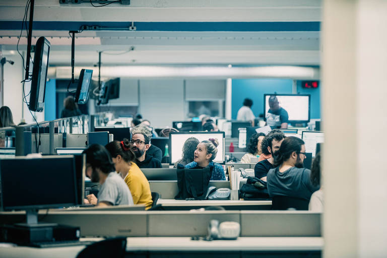 A imagem mostra a Redação da Folha, um ambiente de escritório com várias pessoas trabalhando. Há computadores em cada mesa e algumas pessoas estão interagindo entre si. O espaço é iluminado e parece ser um ambiente colaborativo.