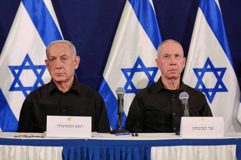 A imagem mostra Netanyahu e Gallant sentados em uma mesa durante uma conferência de imprensa. Os dois estão vestidas com camisas pretas e há bandeiras de Israel ao fundo. As bandeiras têm o símbolo da Estrela de Davi e estão dispostas em um padrão alternado.