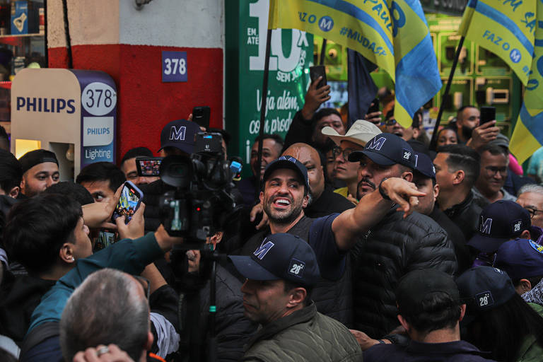 A imagem mostra um grupo de pessoas em uma manifestação, com várias bandeiras azuis e amarelas. Algumas pessoas estão segurando celulares, enquanto outras estão em volta de um homem sorridente que parece estar interagindo com a multidão. Ao fundo, há um letreiro da Philips e um número 378 na parede. A atmosfera é de entusiasmo e apoio.