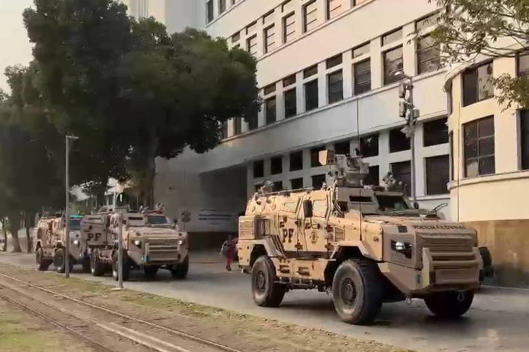 A imagem mostra um grupo de veículos blindados em uma rua, passando em frente a um edifício de aparência institucional. Os veículos são de cor bege e possuem marcas visíveis, incluindo a sigla 'PF'.