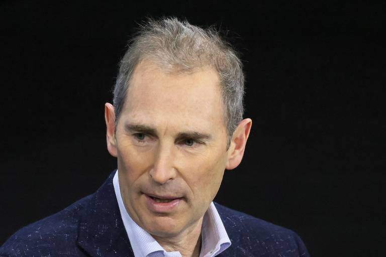 O CEO da Amazon, Andy Jassy, homem branco, com cabelo curto e grisalho, usando um paletó escuro, está em um ambiente com fundo escuro