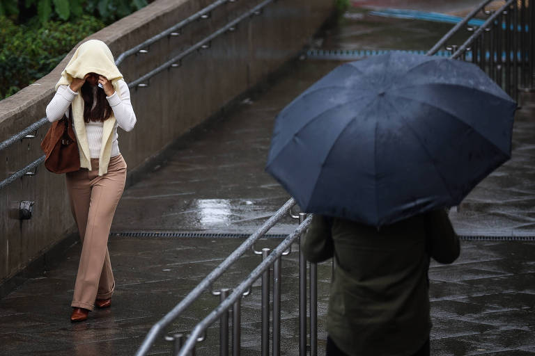 Uma mulher caminha em uma calçada molhada, cobrindo a cabeça com uma blusa clara, enquanto um homem com um guarda-chuva preto está em sua direção. O ambiente parece chuvoso, com reflexos na superfície molhada e vegetação ao fundo.