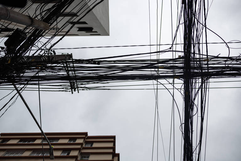 A imagem mostra uma vista de baixo para cima de fios elétricos e de telecomunicações cruzando o céu nublado, formando uma cruz. No fundo, é possível ver a silhueta de um edifício residencial com janelas. O céu está cinza, sugerindo um clima nublado.
