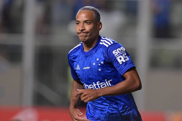 Um jogador de futebol está correndo em campo, vestindo uma camisa azul com detalhes brancos e o número 10. Ele parece estar sorrindo ou fazendo uma expressão de satisfação. Ao fundo, um jogador adversário está caindo no chão, vestindo uma camisa vermelha. O ambiente é um estádio de futebol, com torcedores visíveis nas arquibancadas.