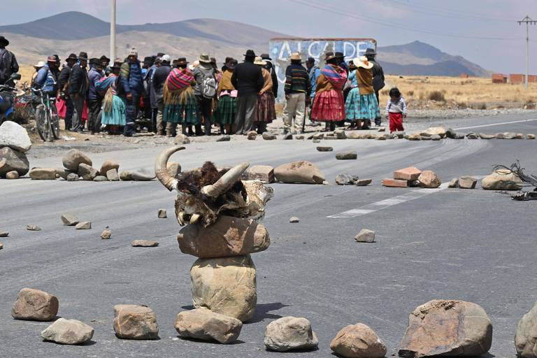 A imagem mostra um grupo de pessoas reunidas ao fundo, em uma estrada bloqueada por pedras. No primeiro plano, há uma grande pedra com chifres de animal, cercada por outras pedras. As pessoas estão vestidas com roupas tradicionais bolivianas. Ao fundo, é possível ver uma placa que diz 'Alcalde'.