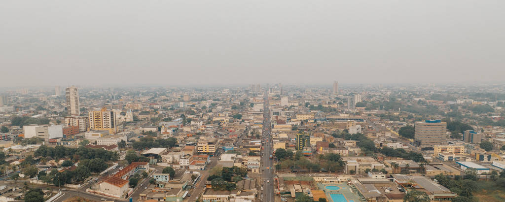 A imagem mostra uma vista aérea de uma cidade com uma grande avenida central. Há diversos edifícios de diferentes tamanhos e estilos ao longo da avenida, além de áreas verdes e algumas construções de telhado vermelho. O céu está nublado, dando um aspecto de neblina à cena.