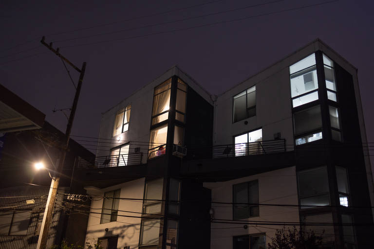 A imagem mostra um edifício moderno à noite, com algumas janelas iluminadas. O prédio tem uma estrutura angular e é composto por paredes brancas e janelas de vidro. À esquerda, há um poste de luz e fios expostos. A iluminação da rua é visível, criando um contraste com o céu escuro.