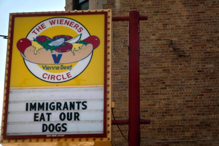 A placa do restaurante The Wieners Circle apresenta um desenho de um cachorro-quente com ingredientes como cebola, picles e molho. Abaixo do desenho, há uma mensagem em letras grandes que diz: 'IMMIGRANTS EAT OUR DOGS'. O fundo da placa é amarelo e a parte inferior é branca. A placa está fixada em um suporte vermelho, com uma parede de tijolos ao fundo.
