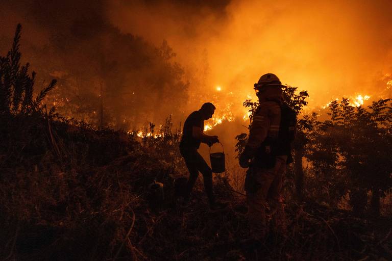 A imagem mostra duas pessoas em uma área afetada por um incêndio florestal. O fundo é dominado por chamas intensas e fumaça, criando uma atmosfera de emergência. Um dos bombeiros está agachado, enquanto o outro está em pé, ambos usando equipamentos de proteção.
