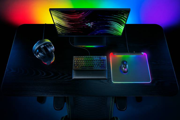 Imagem de um setup de computador gamer visto de cima. Um monitor com luzes coloridas ao fundo, um teclado mecânico iluminado, um mouse com um mousepad iluminado, e um fone de ouvido ao lado. O ambiente é escuro, destacando as luzes RGB dos equipamentos.