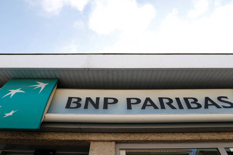 A imagem mostra a fachada de uma agência do banco BNP Paribas. O logotipo do banco é visível, com um fundo verde e estrelas brancas. Abaixo do logotipo, está escrito 'BNP PARIBAS' em letras grandes e escuras. O céu está claro ao fundo.
