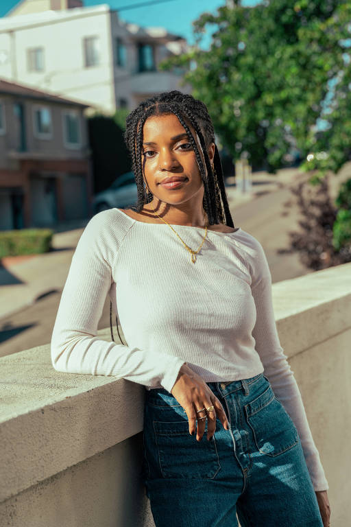 Uma mulher negra jovem está posando ao ar livre, encostada em uma parede. Ela usa uma blusa de manga longa branca e jeans. Ao fundo, há casas e árvores, com um céu claro e ensolarado.