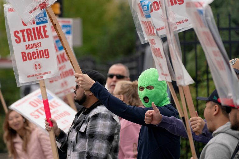 A imagem mostra um grupo de manifestantes segurando cartazes em um protesto contra a Boeing. Um dos manifestantes, usando uma máscara verde, levanta um cartaz que diz 'STRIKE AGAINST Boeing'. Outros manifestantes estão ao fundo, alguns segurando cartazes semelhantes. O ambiente parece ser urbano, com árvores e uma cerca ao fundo.
