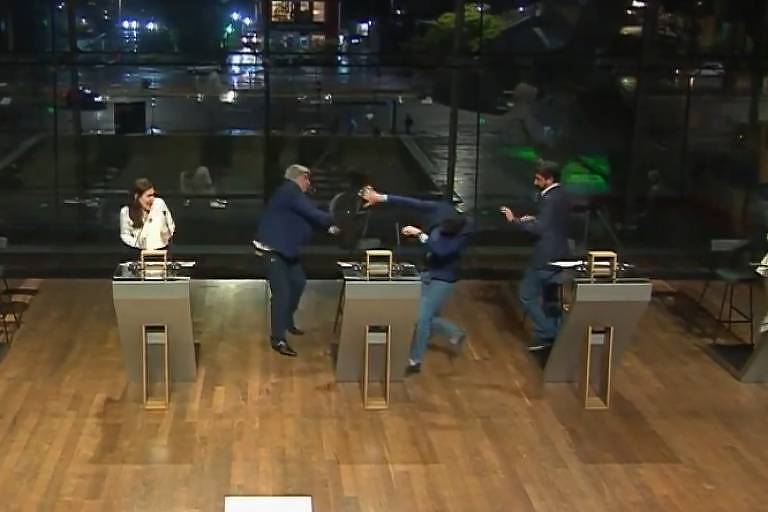 A imagem mostra um estúdio de televisão onde ocorre um debate. Há seis pessoas e duas delas estão em confronto, uma arremessando a cadeira na outra. No canto inferior direito, uma pessoa está fazendo interpretação em Libras. Ao fundo, é possível ver uma cidade iluminada à noite. Na parte inferior da imagem, há um banner com o texto: 'Jornal da Cultura Datena é expulso, Marçal deixa debate'.