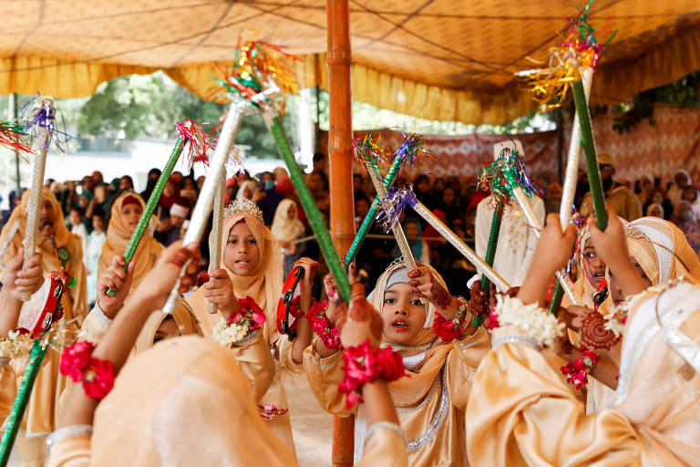 Um grupo de meninas vestidas com roupas douradas e adornos florais participa de uma dança tradicional sob uma tenda. Elas seguram bastões decorados com fitas coloridas e estão em movimento, enquanto uma multidão observa ao fundo. O ambiente é festivo e alegre.