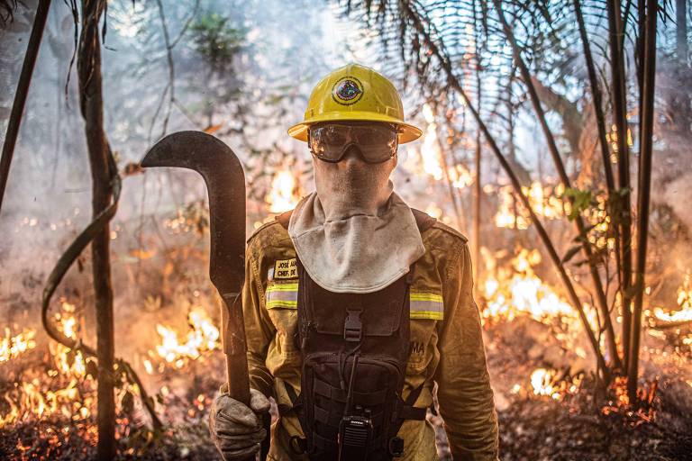 Brigadista usando máscara e foice para combater incêndios com floresta queimando ao fundo