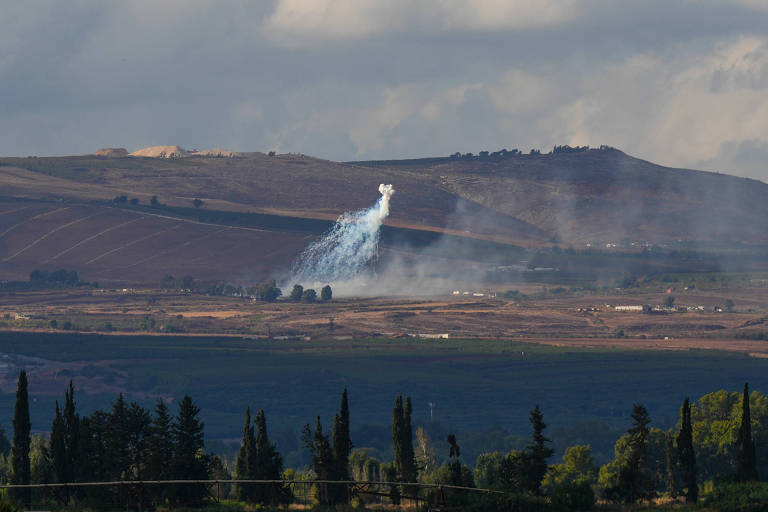 Em um campo com montanha ao fundo, a explosão de projétil de artilharia precipita fragmentos e fumaça ao solo 