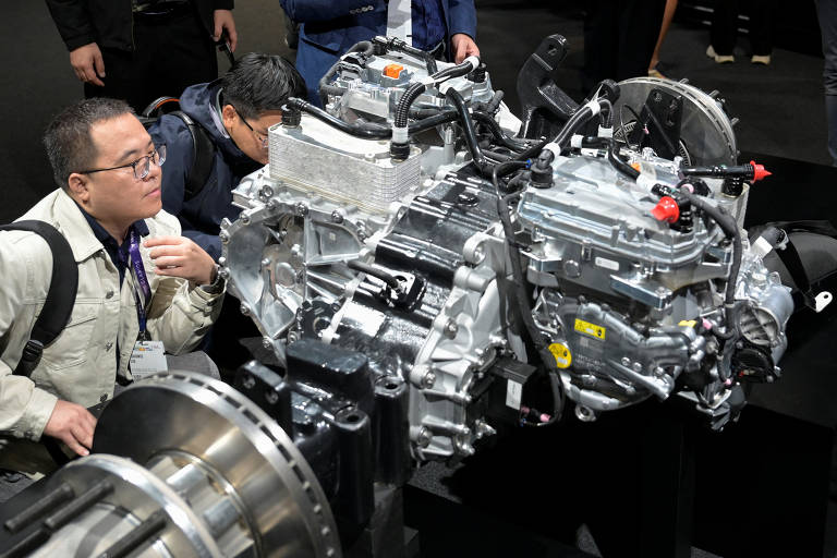 A imagem mostra um motor exposto em uma feira ou evento, com duas pessoas observando de perto. Um homem, com óculos e uma jaqueta clara, está inclinado para examinar o motor, enquanto outro homem, ao fundo, também observa. O motor é detalhado, com várias partes metálicas e fios visíveis, montado sobre uma base preta.