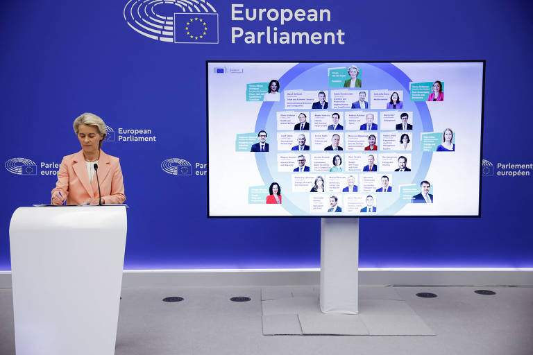 Uma mulher em pé em um púlpito branco, apresentando informações em um grande display digital. O fundo é azul com o logotipo do Parlamento Europeu. O display mostra uma organização ou lista de pessoas, com fotos e nomes dispostos em um formato circular. A mulher está vestindo um blazer claro e parece estar explicando algo ao público.